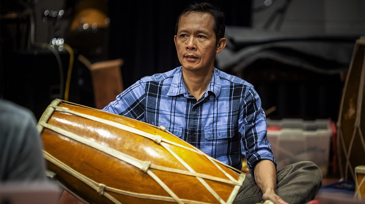 A man in a blue-and-black check shirt sits holding a large two-headed hand drum